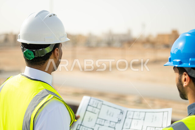 الوقوف في موقع العمل و متابعة سير المشاريع الإعمارية، الاهتمام في بناء الأساسيات الجيده للمباني، التحقق من تطبيق الخطط الهندسيه المعمارية أثناء إنشاء المبنى ,صورة مقربة من الخلف لمهندسان عربيان خليجيان سعوديان يرتديان الخوذة وسترة الحماية يمسكان المخطط الهندسي