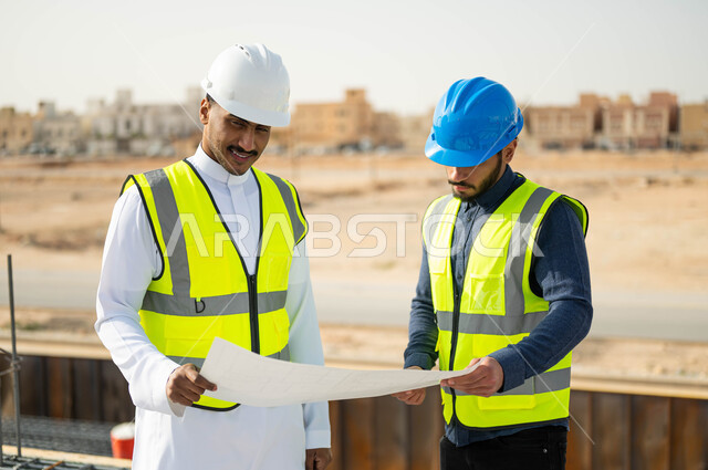 Exchanging ideas and discussions about the work project, interest in building good basics for the building, following up on the progress of construction projects, verifying the correctness of the application of architectural plans in the building, a close-up photo of two Saudi Gulf Arab engineers wearing helmets and protection vests, holding the building plan.