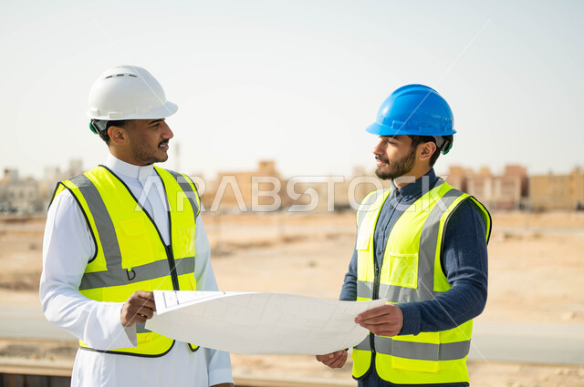 Exchanging ideas and discussions about the work project, interest in building good basics for the building, following up on the progress of construction projects, verifying the correctness of the application of architectural plans in the building, a close-up photo of two Saudi Gulf Arab engineers wearing helmets and protection vests, holding the building plan.