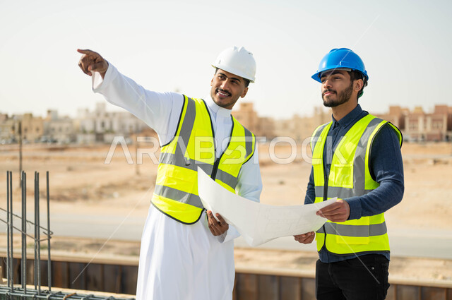 Referring to something, the gestures of conviction and admiration for the progress of architectural projects in the correct way, verify the application of architectural plans during the construction of the building, two Saudi Gulf workers and engineers wearing helmet and protection jacket holds the scheme and exchanges engineering ideas