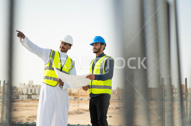 الاشارة إلى شيء ما ،متابعة سير المشاريع الإعمارية, التحقق من صحه تطبيق الخطط الهندسيه المعمارية أثناء إنشاء المبنى ،الحرص على بناء الأساسيات الجيده للمباني، عامل ومهندس عربيان خليجيان سعوديان يرتديان الخوذة وسترة الحماية يمسكان المخطط ويتبادلان الافكار الهندسيه