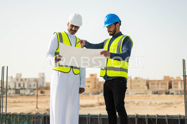الاهتمام في بناء الأساسيات الجيده للمباني ،متابعة سير المشاريع الإعمارية, التحقق من صحه تطبيق الخطط الهندسيه المعمارية أثناء إنشاء المبنى، عامل ومهندس عربيان خليجيان سعوديان يرتديان الخوذة وسترة الحماية في موقع العمل، الاشارة إلى شيء ما