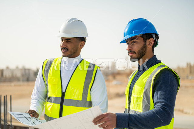 الاهتمام في بناء الأساسيات الجيده للمباني، متابعة سير المشاريع الإعمارية, التحقق من تطبيق الخطط الهندسيه المعمارية أثناء إنشاء المبنى ,صورة مقربة لعامل ومقاول عربيان خليجيان سعوديان يرتديان الخوذة وسترة الحماية يمسكان المخطط ويتبادلان الافكار الهندسيه