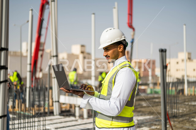 انجاز المهام المطلوبة ، تدوين ملاحظات العمل عبر جهاز الحاسوب المحمول ،رسم المخططات من خلال البرامج الهندسية، العمل في مجال الهندسة المعمارية ،وظائف و مهن سعودية، صورة مقربة لمهندس معماري عربي خليجي سعودي يرتدي الخوذة والسترة الخاصة بالعمل