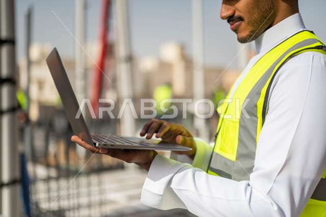 Completing the required tasks, taking work notes via the laptop, drawing plans through engineering programs, working in the field of architecture, Saudi jobs and professions, a close-up picture of a Saudi Gulf Arab architect wearing a helmet and work jacket.