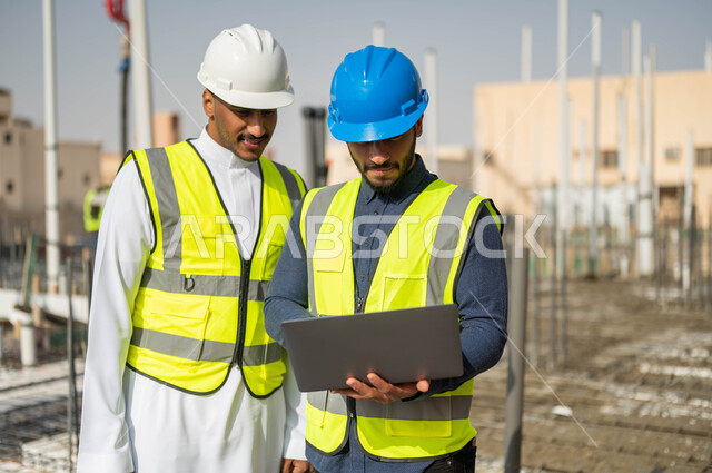 Taking work notes on a laptop, drawing plans using engineering programs, working in the field of architecture, Saudi jobs and professions, a close-up photo of two Saudi Gulf Arab architects wearing helmets and work jackets.