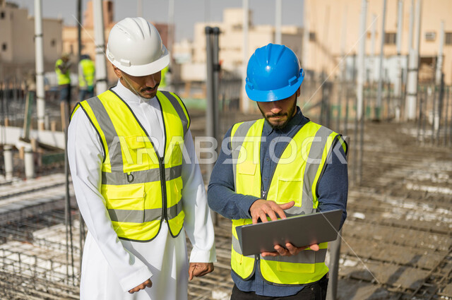 رسم المخططات من خلال البرامج الهندسية، الاطلاع على تصاميم هندسية جديدة عبر جهاز الحاسوب المحمول ، العمل في مجال الهندسة المعمارية ،وظائف و مهن سعودية، صورة مقربة لمهندسان معماريان عربيان خليجيان سعوديان يرتديان الخوذة والسترة الخاصة بالعمل