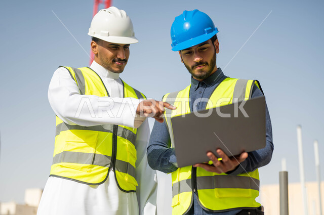 رسم المخططات من خلال البرامج الهندسية، العمل في مجال الهندسة المعمارية ،وظائف و مهن سعودية، الاشارة إلى جهازالحاسوب المحمول و الاطلاع على تصاميم هندسية جديدة ،صورة مقربة لمهندسان معماريان عربيان خليجيان سعوديان يرتديان الخوذة والسترة الخاصة بالعمل