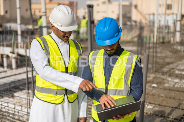 العمل في مجال الهندسة المعمارية ،وظائف و مهن سعودية، الاشارة إلى جهازالحاسوب المحمول و الاطلاع على تصاميم هندسية جديدة، دمج العمل مع التقنية و التكنولوجيا ،صورة مقربة لمهندسان معماريان عربيان خليجيان سعوديان يرتديان الخوذة والسترة الخاصة بالعمل