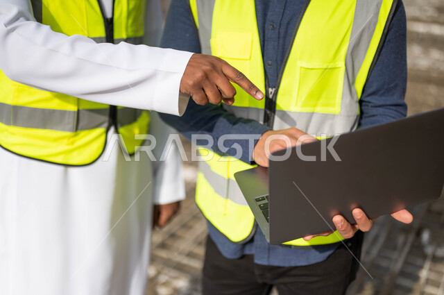 مهن ووظائف سعودية، ارتداء اللباس الخاص بالعمل، الاشارة إلى جهازالحاسوب المحمول و الاطلاع على تصاميم هندسية جديدة، دمج العمل مع التقنية و التكنولوجيا ،صورة مقربة لمهندسان معماريان عربيان خليجيان سعوديان ،العمل في مجال الهندسة المعمارية