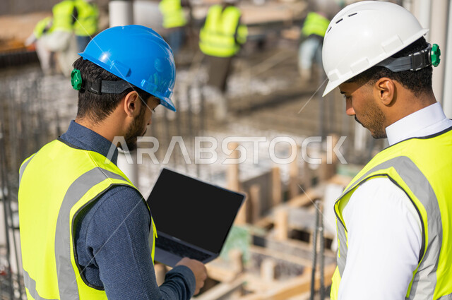 مشاركة الآراء بين الزملاء في الاطلاع على تصاميم هندسية جديدة عبر جهاز الحاسوب المحمول، دمج العمل مع التقنية و التكنولوجيا ،صورة مقربة من الخلف لمهندسان معماريان عربيان خليجيان سعوديان يرتديان الخوذة وسترة الحماية يعملان في مجال العمارة