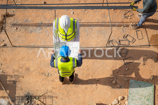Wearing a work jacket, applying engineering projects and plans in Saudi Arabia, exchanging conversations and conversations about the project and pointing to something, an overhead vertical picture of two Saudi Gulf Arab architects wearing helmets working in the field of architecture.