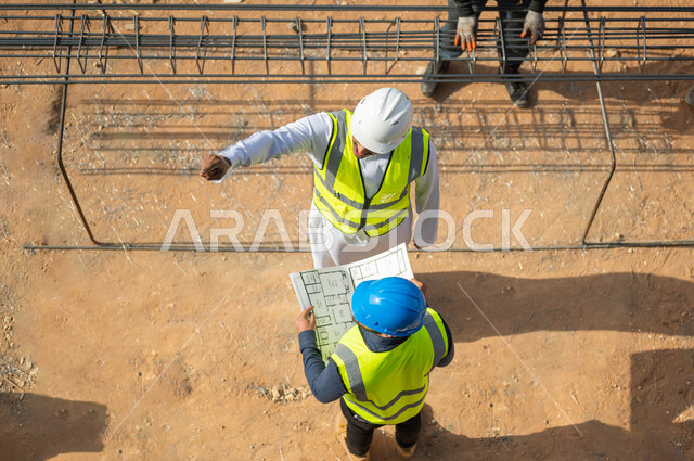 Implementing engineering projects and plans in Saudi Arabia < Paying attention to the quality of raw materials, exchanging conversations and chats about the project and pointing to something, an overhead vertical picture of two Saudi Gulf Arab architects wearing helmets and protection vests working in the field of architecture