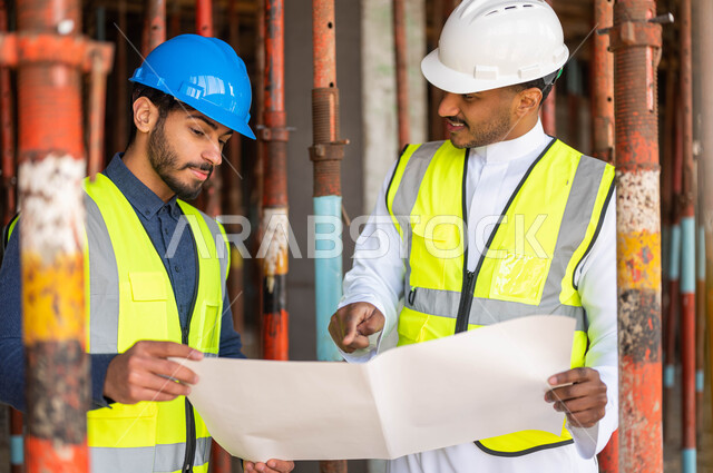 الاشراف على تطبيق المشاريع والخطط الهندسيه في السعودية ،الاشارة إلى مخطط البناء وتبادل الحوارات و الأحاديث التي تدور حول المشروع، صورة مقربة لمهندسان معماريان عربيان خليجيان سعوديان يرتديان الخوذة وسترة الحماية يعملان في مجال الهندسة المعمارية
