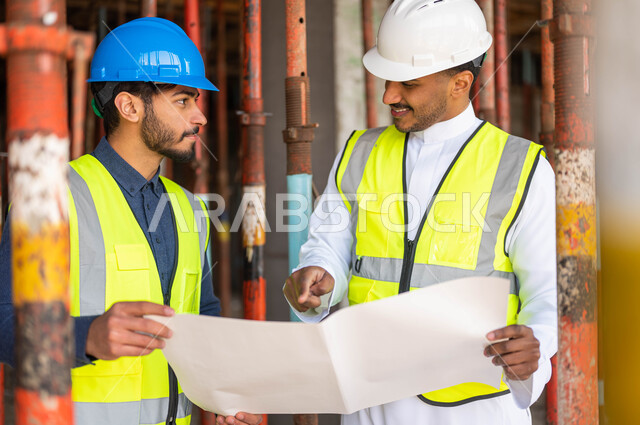 الاشارة إلى مخطط البناء وتبادل الحوارات و الأحاديث التي تدور حول المشروع، الاشراف على تطبيق الخطط و المشاريع الهندسيه في السعودية ،صورة مقربة لمهندسان معماريان عربيان خليجيان سعوديان يرتديان الخوذة وسترة الحماية يعملان في مجال الهندسة المعمارية