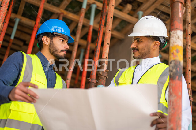 الحوارات و الأحاديث التي تدور حول المشروع بإيماءات السرور، الاشراف على تطبيق الخطط و المشاريع الهندسيه في السعودية ,المساعدة في اقتراح أفكار جديدة ،صورة مقربة لمهندسان معماريان عربيان خليجيان سعوديان يرتديان الخوذة وسترة الحماية يعملان في مجال الهندسة المعمارية