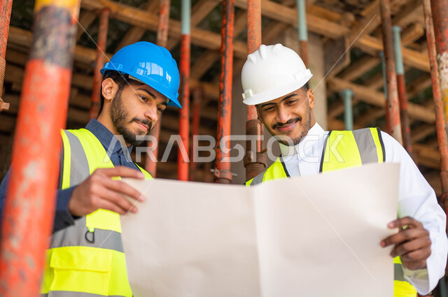 الاشراف على تطبيق الخطط و المشاريع الهندسيه في السعودية ,المساعدة في اقتراح أفكار جديدة ،صورة مقربة لمهندسان معماريان عربيان خليجيان سعوديان يرتديان الخوذة وسترة الحماية يعملان في مجال الهندسة المعمارية، الأحاديث والدردشات التي تدور حول المشروع بإيماءات السرور
