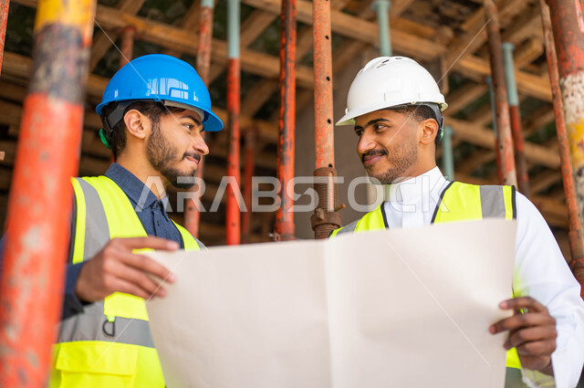 المساعدة في اقتراح أفكار جديدة ،صورة مقربة لمهندسان معماريان عربيان خليجيان سعوديان يرتديان الخوذة وسترة الحماية يعملان في مجال الهندسة المعمارية، الأحاديث والدردشات التي تدور حول المشروع بإيماءات السرور، الاشراف على تطبيق الخطط و المشاريع الهندسيه في السعودية