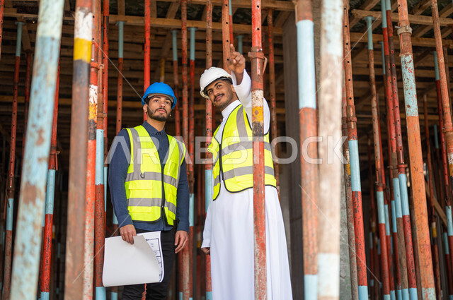 الاشارة الى شيء ما، ايماءات السرور بتبادل الأحاديث والدردشات حول المشروع، عامل ومهندس معماري عربيان خليجيان سعوديان يرتديان الخوذة وسترة الحماية يعملان في مجال الهندسة المعمارية ،الاشراف على تطبيق الخطط و المشاريع الهندسيه في السعودية 