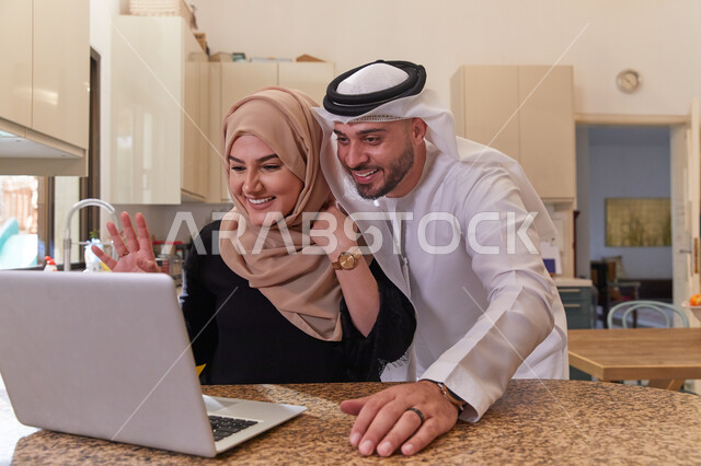 القيام بالأعمال الأونلاين من المنزل، إجراء مكالمات الفيديو والقيام بالبثوث المباشره، زوجان عربيان خليجيان اماراتيان يقومان بالتواصل مع الأصدقاء عبر جهاز الحاسوب المحمول، ايماءات وجه ويدين مختلفة تدل على الفرح والسعاده.