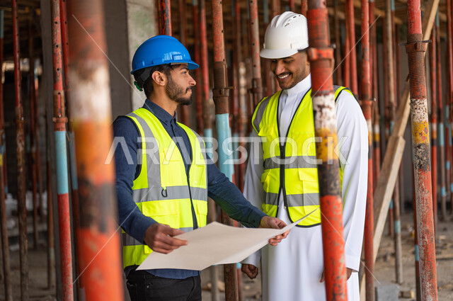 ايماءات السرور بتبادل الأحاديث والدردشات حول المشروع، عامل ومهندس معماري عربيان خليجيان سعوديان يرتديان الخوذة وسترة الحماية يعملان في مجال الهندسة المعمارية ،الاشراف على تطبيق الخطط و المشاريع الهندسيه في السعودية ، الحرص على وضع الأساسيات الجيده في المبنى