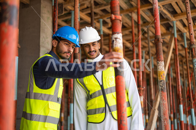 Checking on the raw materials for construction, two Saudi Gulf Arab workers and architects wearing helmets and protective vests working in the field of architecture, supervising the implementation of engineering plans and projects in Saudi Arabia, making sure to put good basics in the building.