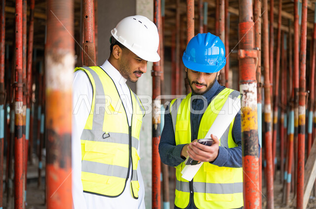 دمج العمل مع التكنولوجيا والتقنية ،مشاركة الرأي مع الزملاء في الاطلاع على تصاميم هندسية جديدة عبر الجوال، عامل ومهندس معماري عربيان خليجيان سعوديان يرتديان الخوذة وسترة الحماية يعملان في مجال الهندسة المعمارية ،النمو والتطور العمراني على أيدي أبناء الوطن