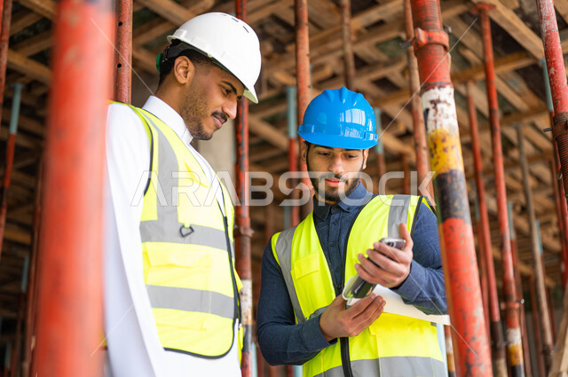 Integrating work with technology and technology, sharing opinions with colleagues in looking at new engineering designs via mobile phone, two Saudi Gulf Arab workers and architects wearing helmets and protective vests working in the field of architecture, urban growth and development at the hands of the people of the country.