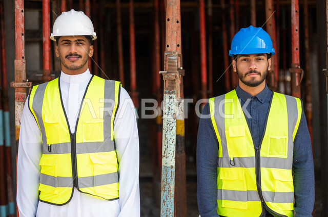 الوقوف باستقامة في موقع العمل ،مواطنان عربيان خليجيان سعوديان يرتديان الخوذة وسترة الحماية  يعملان في مجال الهندسة المعمارية و ينظران إلى الكاميرا ،النمو والتطور العمراني على أيدي أبناء الوطن، الخطط و المشاريع الهندسية