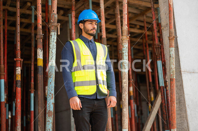 Development and urban growth at the hands of the people of the country, looking at something, taking care to build good basics for the building, applying architectural plans and projects at the work site, a Saudi Gulf Arab engineer wearing a helmet and a protective vest, standing upright and working in the engineering sector.