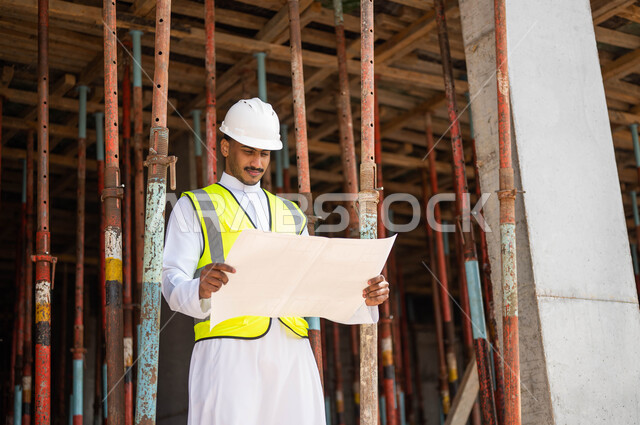 النظر إلى مخطط البناء، الحرص على بناء الأساسيات الجيده للمبنى ،التحقق من تطبيق الخطط و المشاريع الهندسيه المعمارية في موقع العمل ،مهندس عربي خليجي سعودي يرتدي الخوذة وسترة الحماية يعمل في القطاع الهندسي 