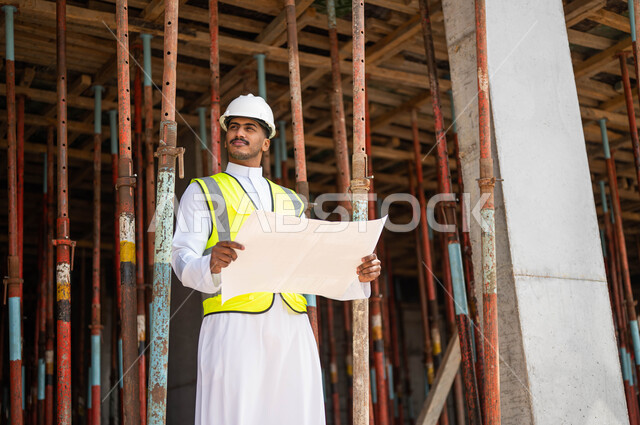 الحرص على بناء الأساسيات الجيده للمبنى، العمل في القطاع الهندسي ،التحقق من تطبيق الخطط و المشاريع الهندسيه المعمارية في البناء, مهندس عربي خليجي سعودي يرتدي الخوذة وسترة الحماية يحمل بيده مخطط وبنظر إلى شيء ما