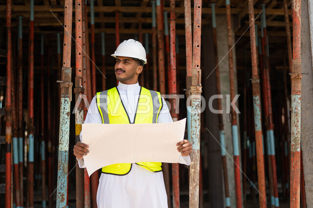 العمل في القطاع الهندسي ،التحقق من تطبيق الخطط و المشاريع الهندسيه المعمارية في البناء بشكل صحيح في السعودية ،الحرص على بناء الأساسيات الجيده للمبنى، صورة مقربة لمهندس عربي خليجي سعودي يرتدي الخوذة وسترة الحماية يحمل بيده مخطط وبنظر إلى شيء ما