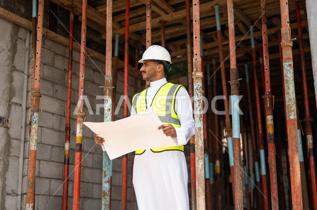 التحقق من تطبيق المشاريع والخطط الهندسيه المعمارية في البناء بشكل صحيح في السعودية ،الحرص على بناء الأساسيات الجيده للمبنى، صورة مقربة لمهندس عربي خليجي سعودي يرتدي الخوذة وسترة الحماية يحمل بيده مخطط وبنظر إلى شيء ما، العمل في القطاع الهندسي