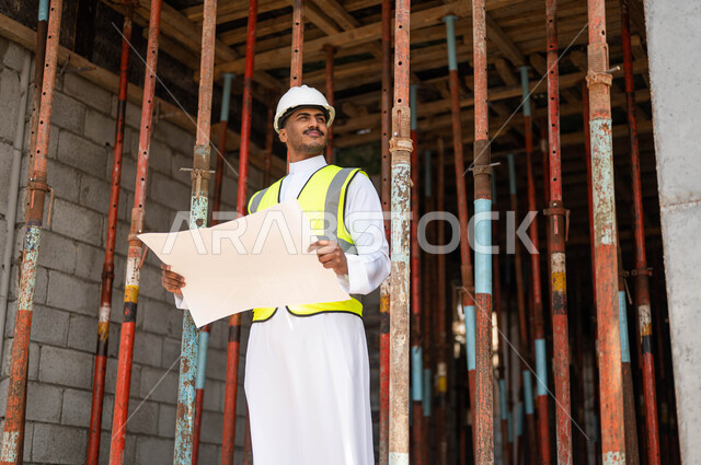 مفهوم الهندسة والانشاءات، الاشراف على تطبيق الخطط و المشاريع الهندسيه المعمارية في السعودية ،الحرص على وضع الأساسيات الجيده في المبنى، مهندس عربي خليجي سعودي مبتسم يرتدي الخوذة وسترة الحماية يحمل بيده مخطط البناء وينظر إلى شيء ما، العمل في القطاع الهندسي
