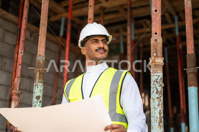 الاشراف على تطبيق الخطط و المشاريع الهندسية المعمارية في السعودية ،الحرص على وضع الأساسيات الجيده في المبنى، العمل في القطاع الهندسي ،مفهوم الهندسة والانشاءات، مهندس عربي خليجي سعودي مبتسم يرتدي الخوذة وسترة الحماية يحمل بيده مخطط البناء وينظر إلى شيء ما