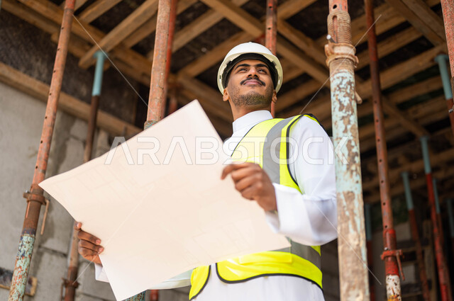Be sure to lay the good basics in the building, work in the engineering sector, the concept of engineering and construction, overseeing the application of architectural plans and projects in Saudi Arabia, a smiling Saudi Arab Gulf engineer who wears the helmet and the protection jacket carries in his hand the construction scheme and looks at something