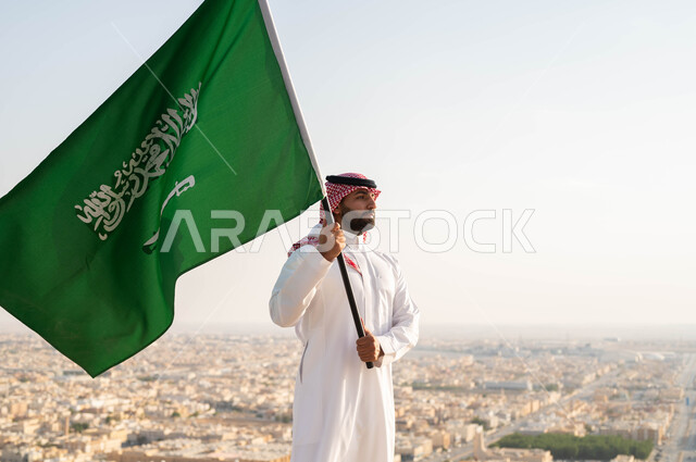 ايماءات الفخر والاعتزاز، الاحتفال بذكرى اليوم الوطني السعودي 23 سبتمبر، صورة مقربة لرجل عربي خليجي سعودي يرتدي الشماغ والزي التقليدي يحمل علم المملكة في العيد الوطني في وضح النهار، يوم العلم 11 مارس، أعياد ومناسبات وطنية ، خلفية أحياء ومباني مدينة الرياض