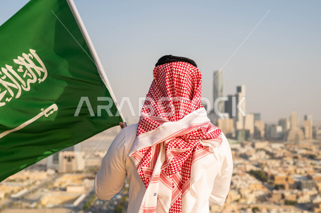 اظهار التفاني للوطن ،الاحتفال بيوم العلم 11 مارس، الأعياد الشعبيه الوطنية في 8 ربيع الاول، إحياء ذكرى اليوم الوطني السعودي 23 سبتمبر، صورة مقربة من الخلف لرجل عربي خليجي سعودي يرتدي الشماغ والزي التقليدي يحمل بيده علم المملكه، خلفية أحياء وشوارع مدينة الرياض.
