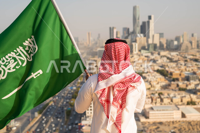 الاحتفال بيوم العلم 11 مارس، الأعياد الشعبيه الوطنية في 8 ربيع الاول، الولاء والاخلاص للوطن، إحياء ذكرى اليوم الوطني السعودي 23 سبتمبر، صورة من الخلف لرجل عربي خليجي سعودي يرتدي الشماغ والزي التقليدي يحمل بيده علم المملكه، خلفية أحياء وشوارع مدينة الرياض.