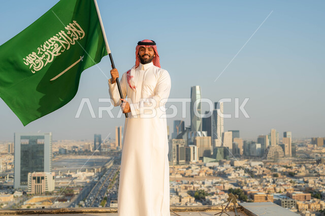 الولاء والاخلاص للبلاد، أعياد ومناسبات وطنية ، يوم العلم 11 مارس، الاحتفال بذكرى اليوم الوطني السعودي 23 سبتمبر، رجل عربي خليجي سعودي مبتسم يرتدي الشماغ والزي التقليدي في العيد الوطني يحمل  بيده علم المملكة في وضح النهار، خلفية الابراج وناطحات السحاب في الرياض