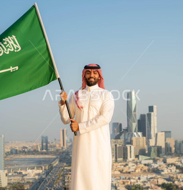 الاخلاص والولاء للبلاد، أعياد ومناسبات وطنية ، يوم العلم 11 مارس، الاحتفال بذكرى اليوم الوطني السعودي 23 سبتمبر، رجل عربي خليجي سعودي مبتسم يرتدي الشماغ والزي التقليدي في العيد الوطني يحمل  بيده علم المملكة في وضح النهار، خلفية الابراج وناطحات السحاب في الرياض