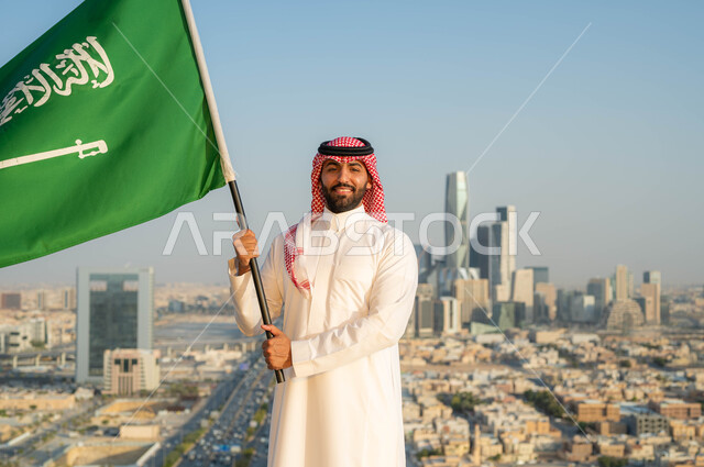 الاخلاص والولاء للبلاد، أعياد ومناسبات وطنية ، يوم العلم 11 مارس، الاحتفال بذكرى اليوم الوطني السعودي 23 سبتمبر، رجل عربي خليجي سعودي مبتسم يرتدي الشماغ والزي التقليدي في العيد الوطني يحمل  بيده علم المملكة في وضح النهار، خلفية الابراج وناطحات السحاب في الرياض