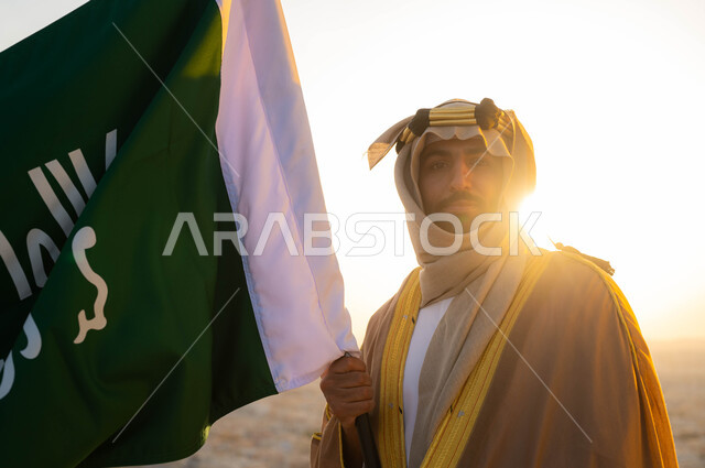 النظر إلى الكاميرا بإيماءات الفخر و الولاء ،ذكرى يوم التأسيس في 22 شباط، توحيد البلاد في 1727 م، الزي الشعبي التقليدي في المنطقة الوسطى ، مؤسس الدولة السعودية الاولى الامام محمد بن سعود, صورة مقربة لرجل عربي خليجي سعودي يرتدي البشت والغترة يحمل بيده علم التأسيس