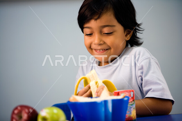 The importance of teaching children the basics of healthy and proper food. A close-up of an Emirati Arab Gulf boy wearing a casual dress and eating a piece of healthy fruit. Looking at something. Enjoying school break time in an Emirati school.