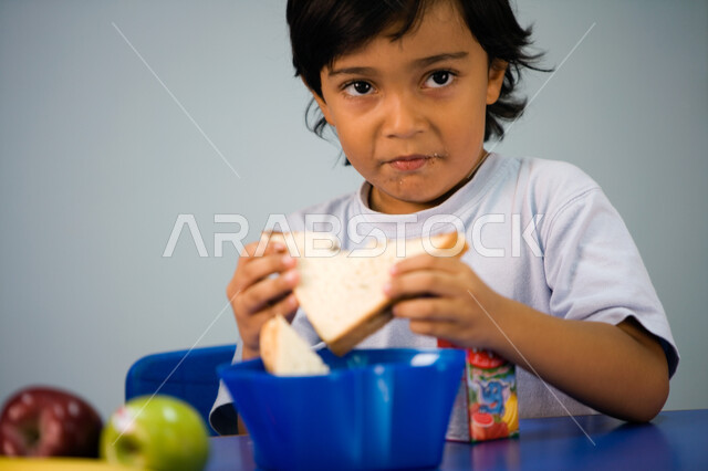 النظر إلى شيء ما بإيماءات التعجب والاستفهام, تعليم الأولاد في الروضة أسس الغذاء الصحي السليم، صورة مقربة لطفل عربي خليجي إماراتي يرتدي الزي الكاجوال, تناول الأطعمة الصحية في وقت الفسحة المدرسية في إحدى المدارس الاماراتية