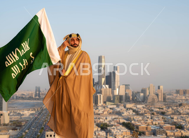 The popular traditional dress in the central region, the celebration of the anniversary of the founding day on February 22, the looks of pride and pride in belonging to the homeland, the unification of the country in 1727 AD, the founder of the first Saudi state, Imam Muhammad bin Saud, a Saudi Gulf Arab man wearing the bisht and ghutra, holding in his hand the founding flag.
