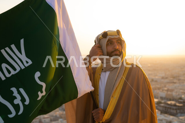 نظرات الفخر و الاعتزاز بالانتماء للوطن، توحيد البلاد في 1727 م، مؤسس الدولة السعودية الاولى الامام محمد بن سعود, صورة مقربة لرجل عربي خليجي سعودي يرتدي البشت والغترة يحمل بيده علم التأسيس, الزي الشعبي في المنطقة الوسطى, الإحتفال بذكرى يوم التأسيس في 22 فبراير