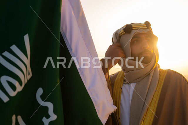 نظرات الاعتزاز والفخر، توحيد البلاد في 1727 م، مؤسس الدولة السعودية الاولى الامام محمد بن سعود, صورة مقربة لرجل عربي خليجي سعودي يرتدي البشت والغترة يحمل بيده علم التأسيس, الزي الشعبي التقليدي في المنطقة الوسطى, الإحتفال بذكرى يوم التأسيس في 22 فبراير