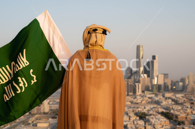 الزي التقليدي ليوم التأسيس, مؤسس الدولة السعودية الاولى الامام محمد بن سعود, ذكرى يوم التأسيس في 22 فبراير، توحيد البلاد وتأسيس السعودية في 1727م, صورة من الخلف لرجل عربي خليجي سعودي يرتدي البشت والغترة يحمل بيده علم التأسيس, خلفية احياء مدينة الرياض.
